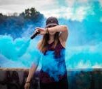 A stylish woman surrounded by bright blue smoke outdoors, creating a dynamic fashion statement.